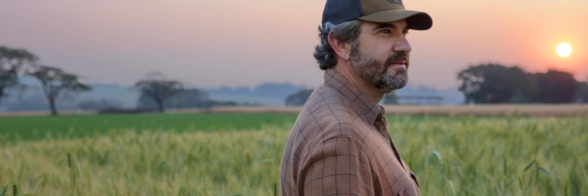 A man in a plaid shirt and baseball cap stands in a field during sunset, gazing to the side.