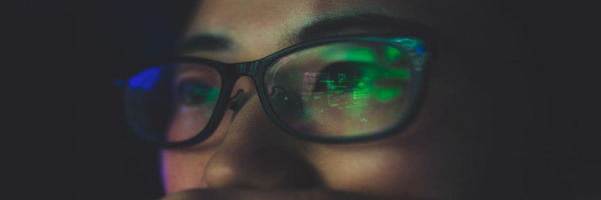 Close-up of a person wearing glasses with computer code reflected on the lenses, suggesting focus on a digital screen in a dimly lit environment.